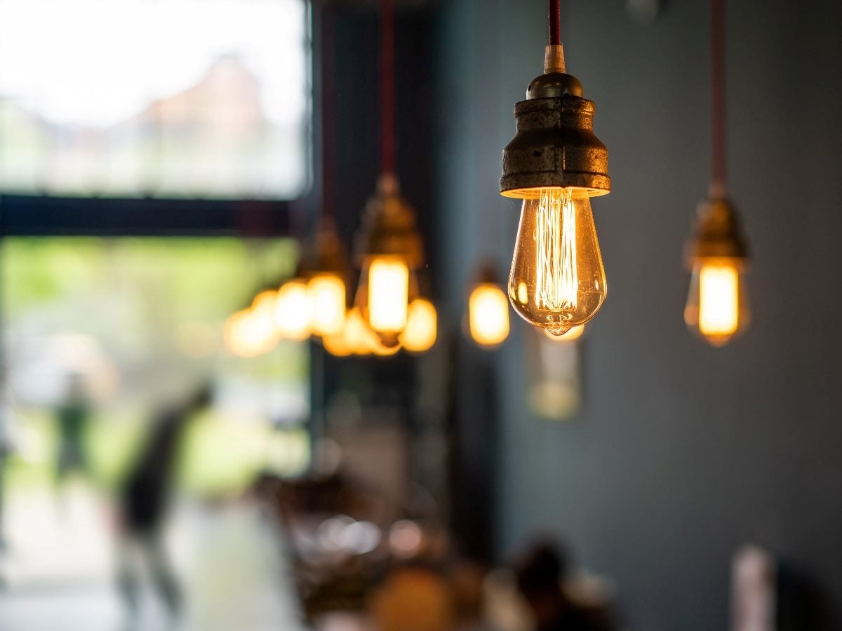 Close-up of a glowing Edison-style light bulb hanging from a cord indoors, with several similar bulbs softly blurred in the background and indistinct figures visible through a window, creating a warm, ambient atmosphere.