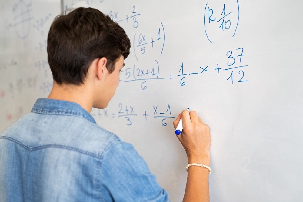 A college student with dark hair and wearing a blue shirt solves an equation on a whiteboard in piece about martech solutions.