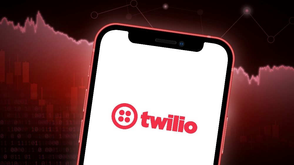 The image features a smartphone centered in the foreground with the Twilio logo displayed on its screen. The background is a dramatic red graphic depicting a fluctuating stock market chart, suggesting a connection between Twilio and market dynamics. The overall color theme is a blend of dark and red hues, creating a sense of urgency or intensity related to the business environment in piece about Twilio.