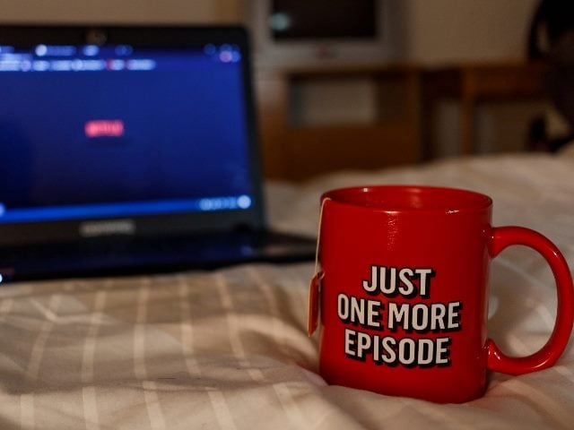 A red coffee mug that says, "Just One More Episode," with a laptop in the background on a bed. The age of ecommerce has changed with viewers of streaming apps being able to purchase products right from their favorite shows.