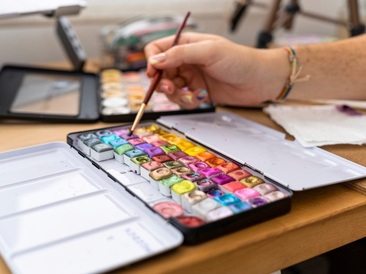 A person’s hand dips a paintbrush into a vibrant watercolor palette filled with colorful paints on a desk, suggesting creativity and artistic expression.