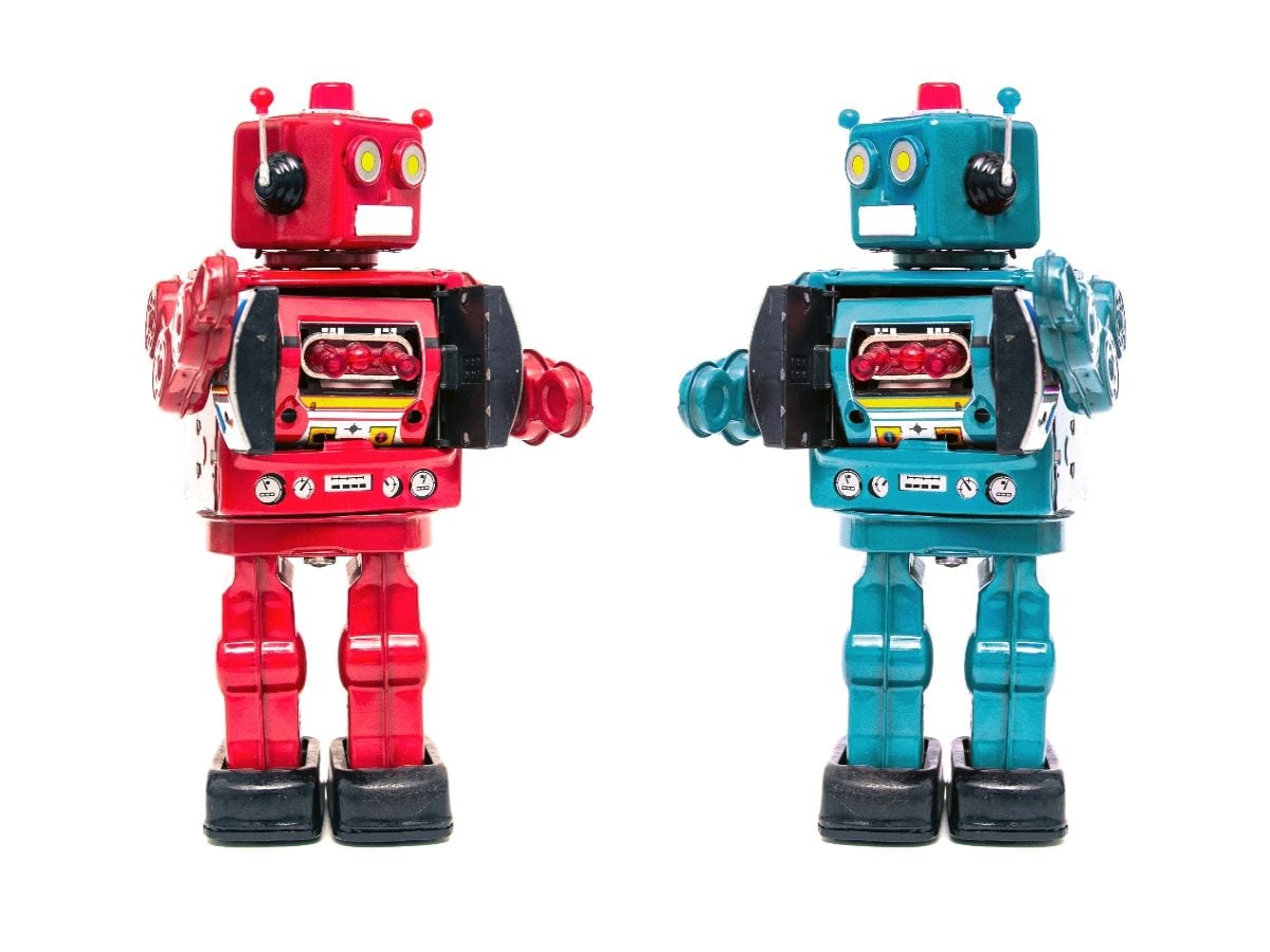 Two vintage-style toy robots, one red and one teal, standing side by side against a plain white background, facing forward with mechanical details and antennae visible.