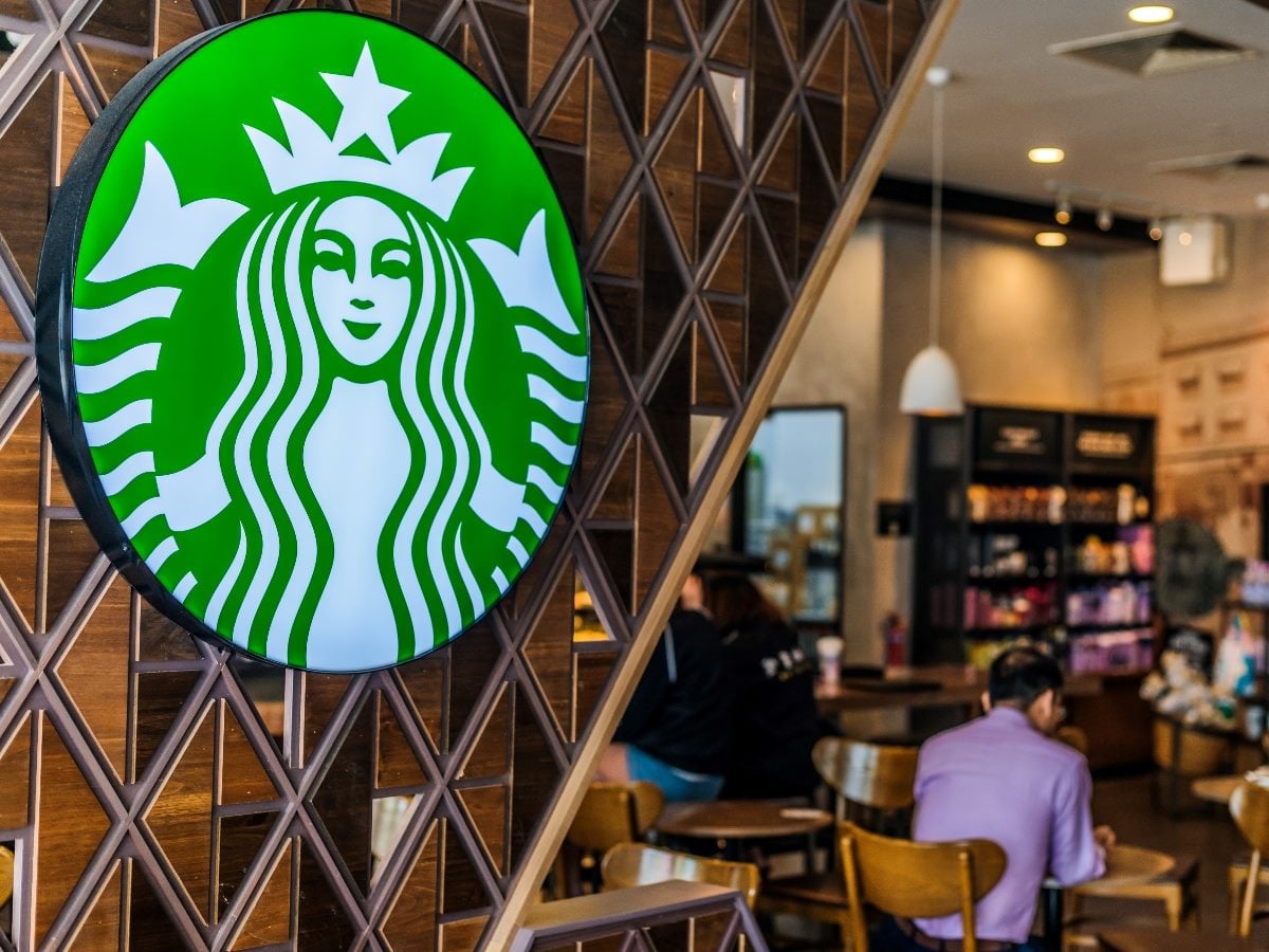 Starbucks siren logo sign on a patterned wall inside a coffee shop, with customers seated at tables in the background.