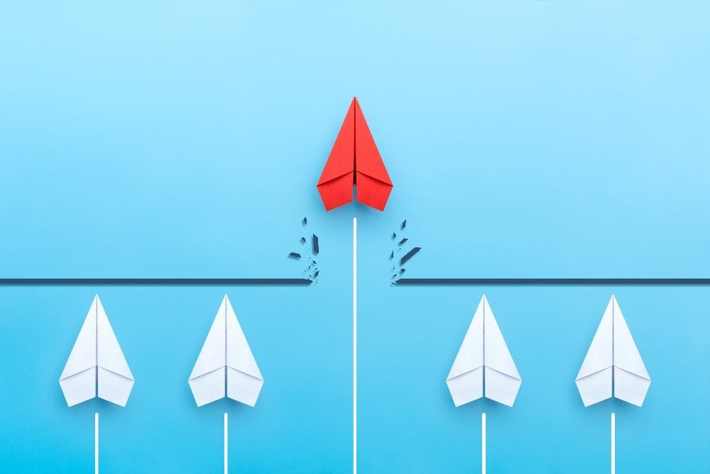 This image depicts several white paper airplanes in a row, all aligned and pointing to the right on a light blue background. One red paper airplane leads the pack, soaring ahead and leaving a trail of small black paper fragments, indicating its rapid movement and breaking away from the standard path set by the others. The scene represents leadership, innovation, and breaking the norm in a visually simple yet powerful way in piece about customer experience strategies.