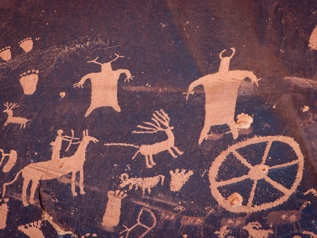 Petroglyphs from Newspaper Rock Historical Site in Utah, light petroglyphs on a dark background depicting various figures and wheels, animals and human foot and hand prints. 
