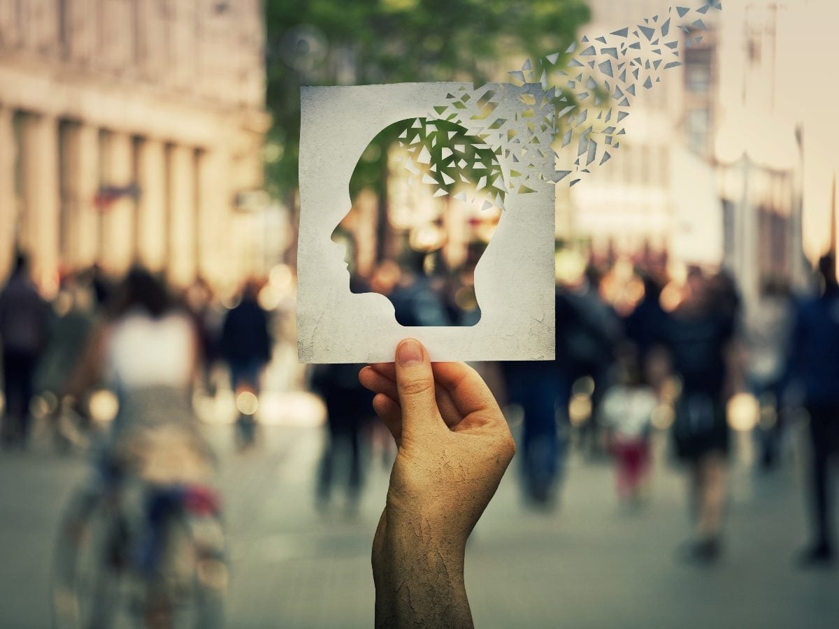 A hand holds a white card with the silhouette of a human head cut out, while small fragmented pieces appear to break away from the top of the head shape. The background shows a blurred city street with pedestrians walking, symbolizing memory loss or fragmented decision-making amid everyday activity.