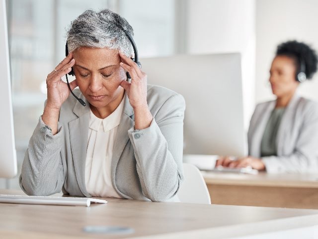 Tired call center woman with headache, with colleague working in the background. 
