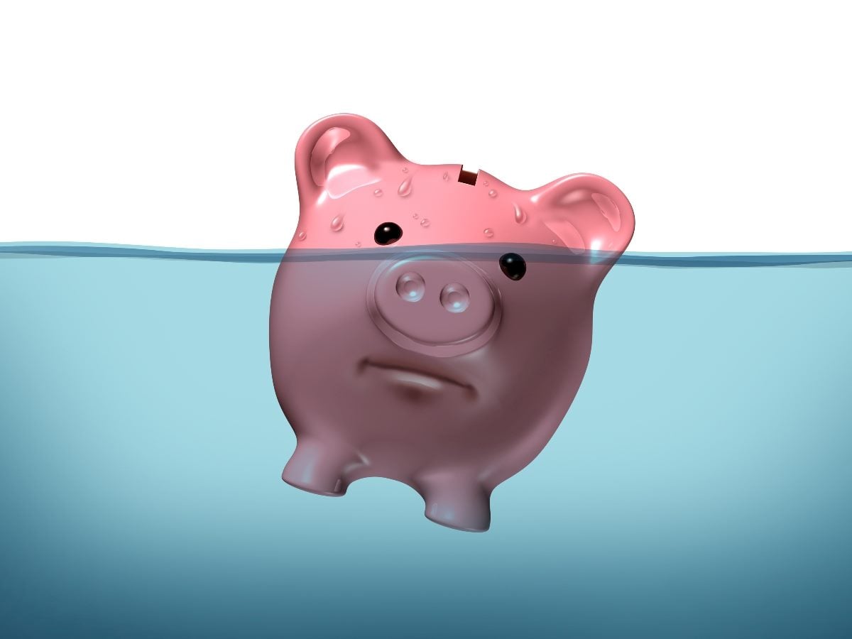 Illustration of a pink piggy bank partially submerged in water, with only the upper portion visible above the surface. The piggy bank has a sad expression, visually suggesting financial stress, debt, sinking finances or economic pressure.