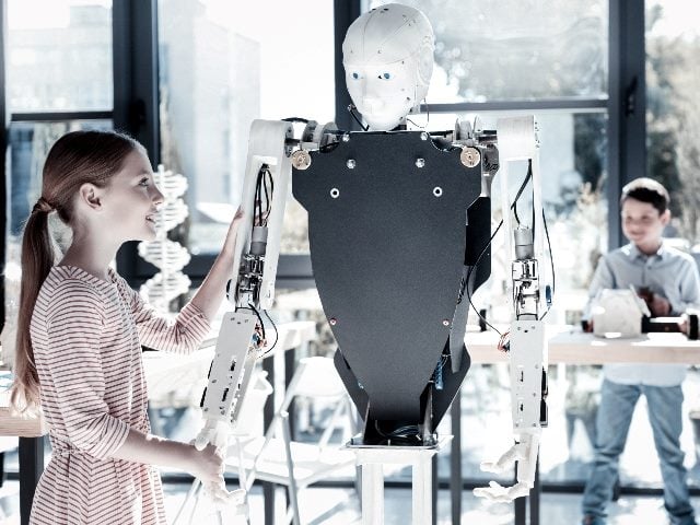 Young lady smiling while holding a hand of a robotic machine and looking at it with excitement as a young boy looks on in the background.