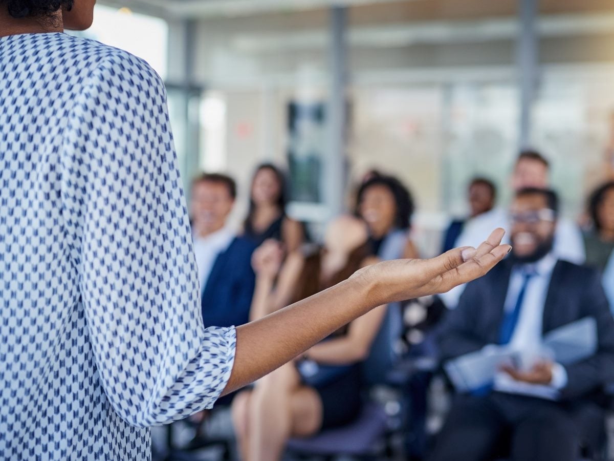 Person speaking to an audience in a conference room, gesturing with one hand while attendees sit blurred in the background.