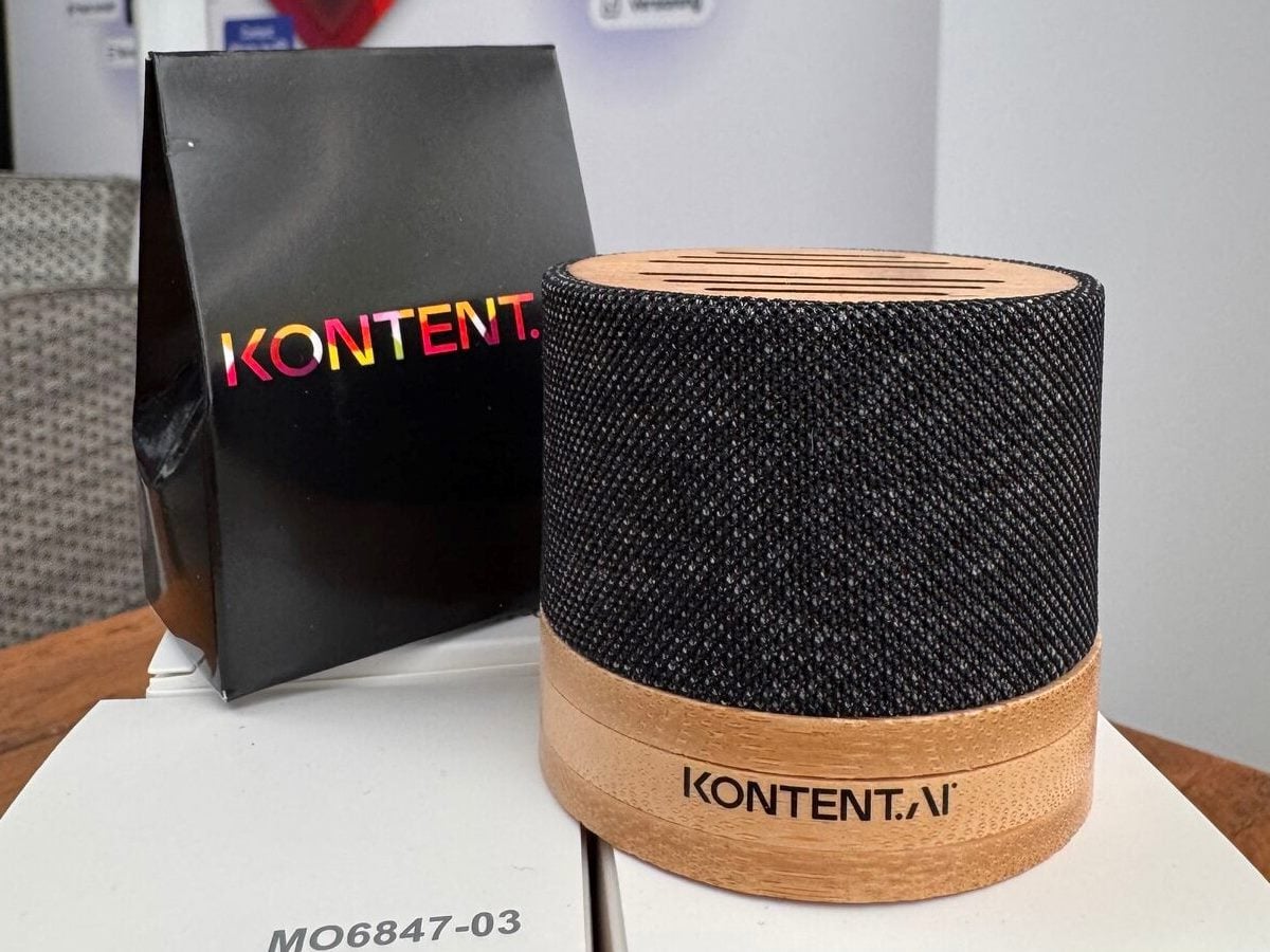Branded Kontent.ai promotional items, including a black gift bag with a colorful Kontent logo and a small cylindrical speaker with a wood base and fabric top printed with “Kontent.ai,” displayed on a desk.