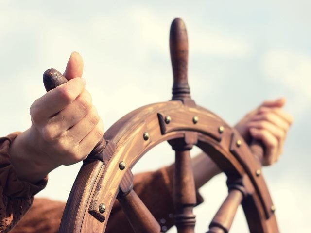 Two hands guide the wooden helm of a boat with the sky in the background, representing the need for a proactive customer experience approach. 