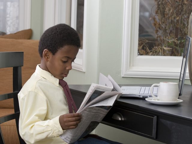 A young boy in a shirt and tie sits down and reads a newspaper.