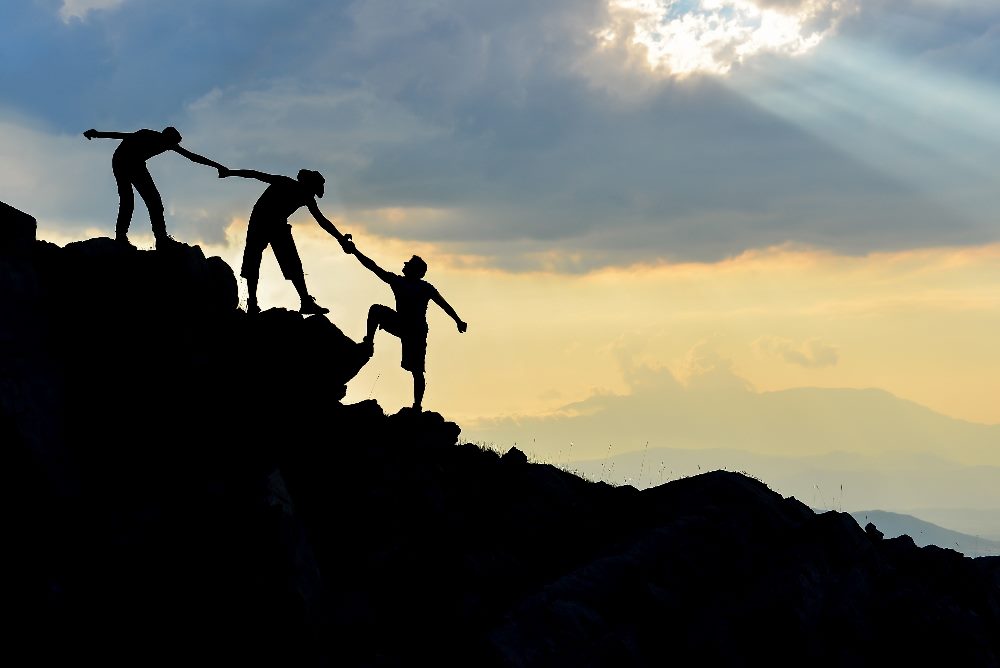 Three people climbing on an incline outdoors link hands and help each other up in piece about CX and EX synergy.