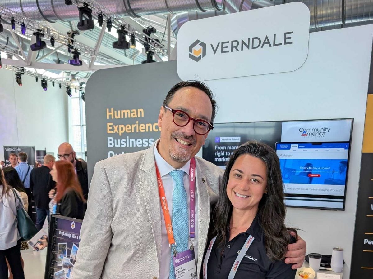 Two professionals smiling and posing together at a Verndale trade show booth, with Verndale branding, digital screens, and a busy conference floor visible in the background.