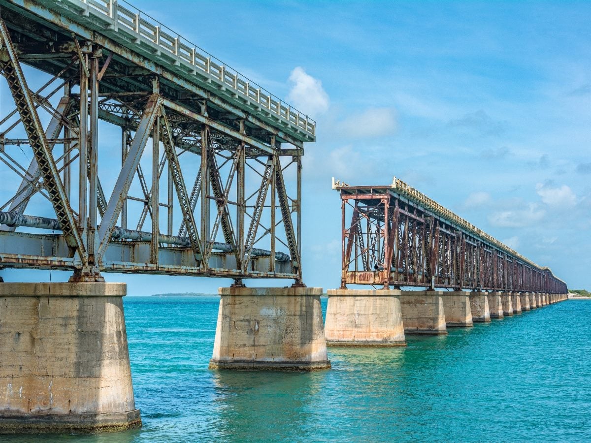 Old and new sections of the Florida Keys Overseas Railroad bridge stretch across turquoise ocean water on concrete piers under a bright blue sky.