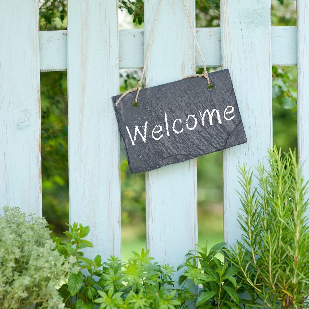 A white picket fence with green plants growing at the bottom has a small slate sign with the word "welcome" on it in a piece about CX and EX journeys.