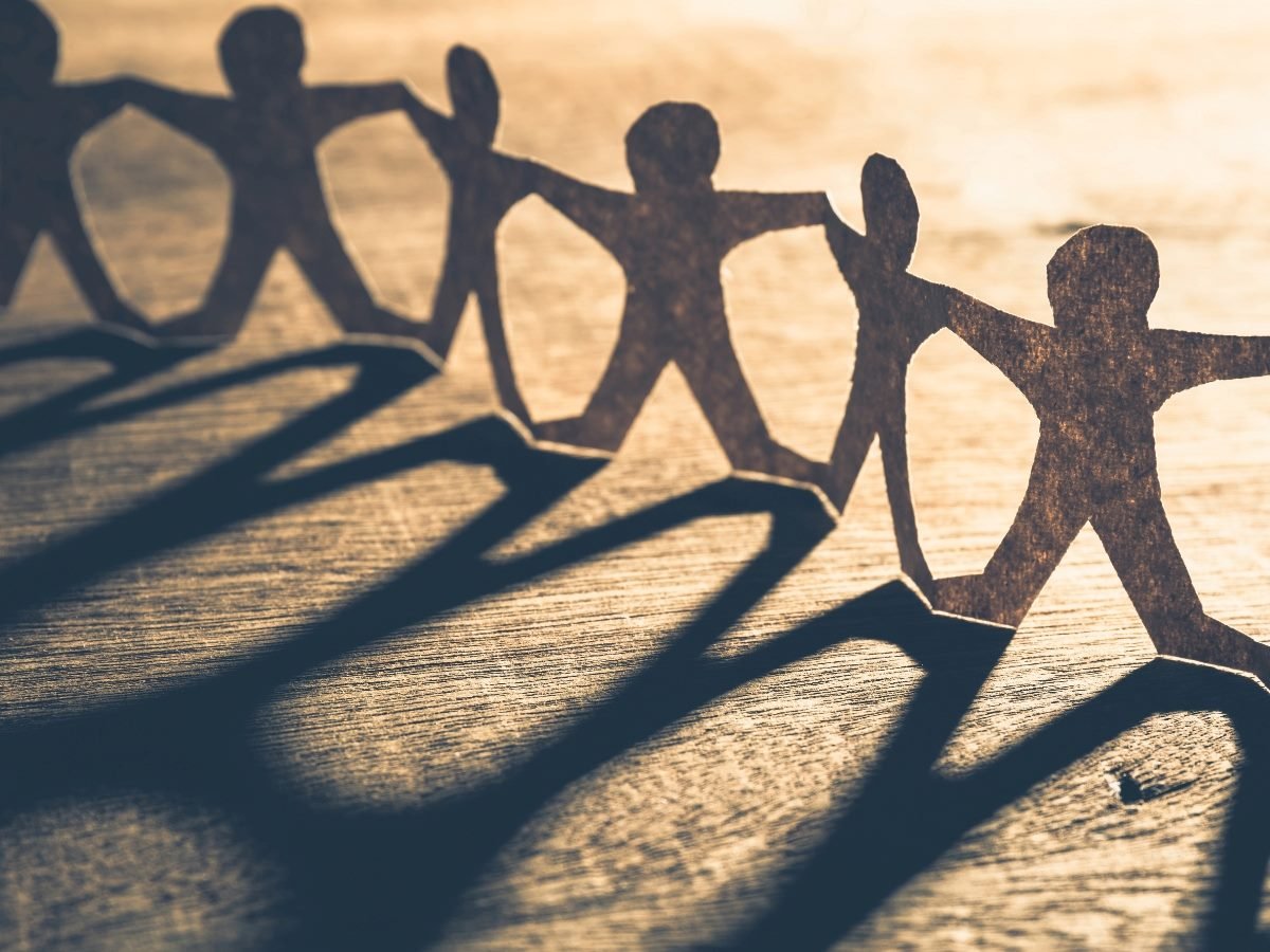 Paper cutout chain of people holding hands with shadows on a wooden surface, symbolizing teamwork, collaboration and connected customer experience.