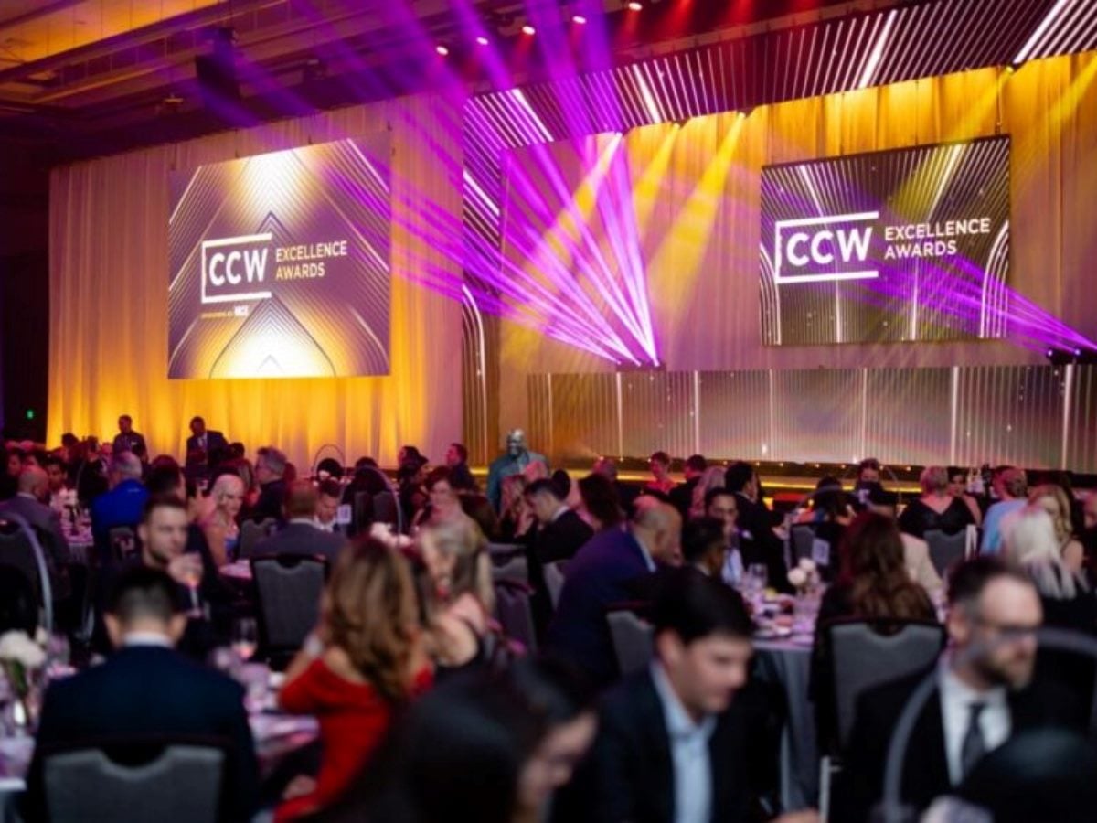 Attendees gather inside a ballroom during the CCW Excellence Awards at Customer Contact Week in Las Vegas. Purple and gold stage lighting illuminates the room while large screens display CCW Excellence Awards branding above seated guests at banquet tables.