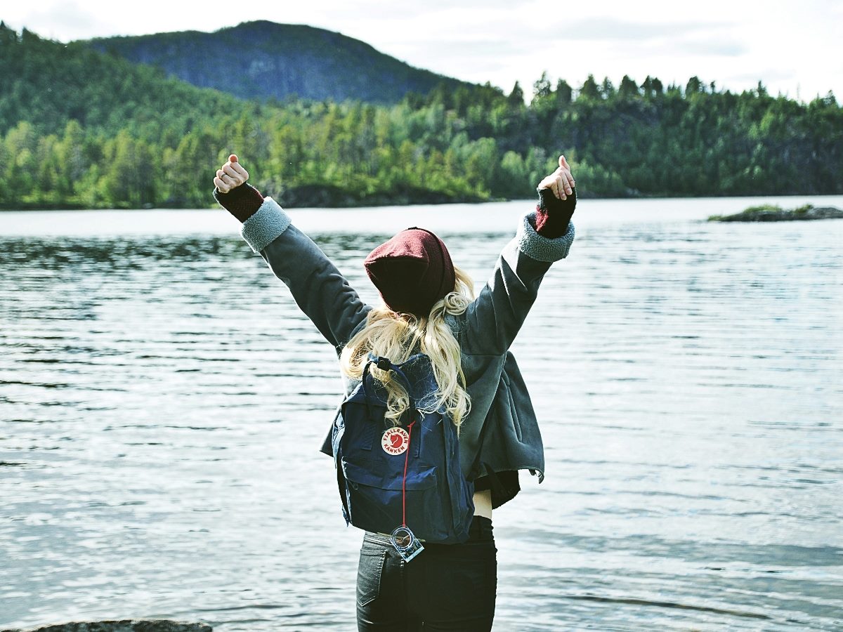 Woman facing a body of water with arms raised.