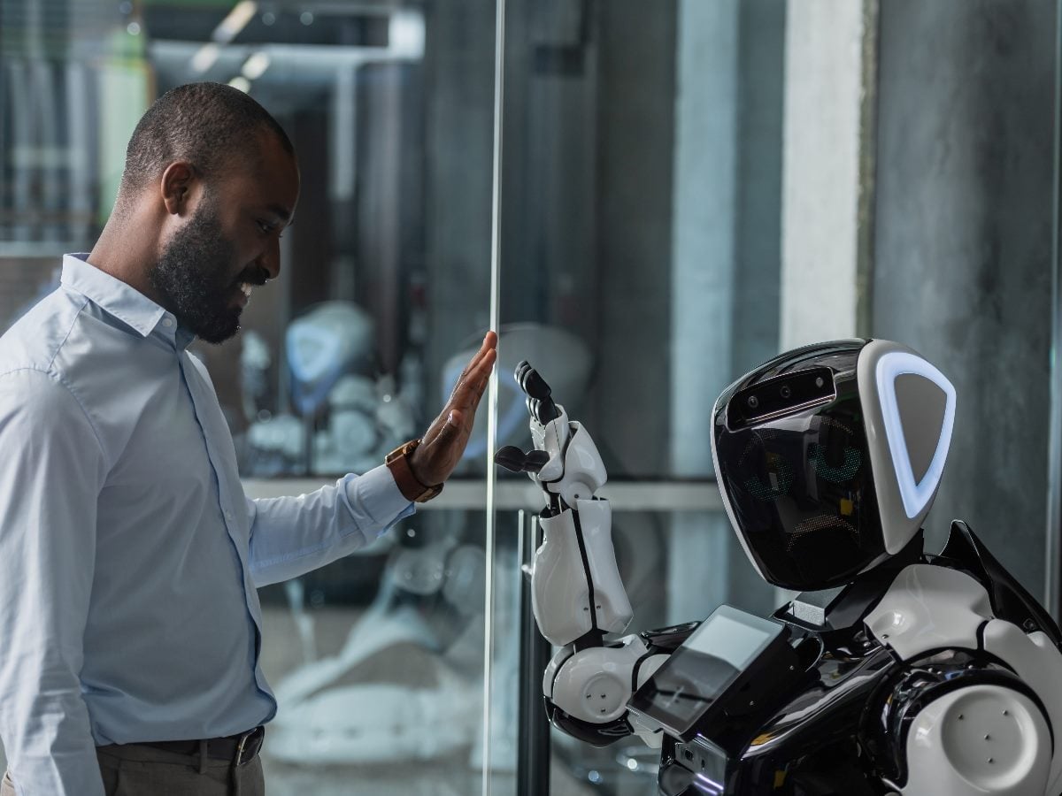 A man smiles as he touches his hand to a glass wall while a humanoid robot on the other side mirrors the gesture, creating a moment of human–AI interaction in a modern indoor setting.