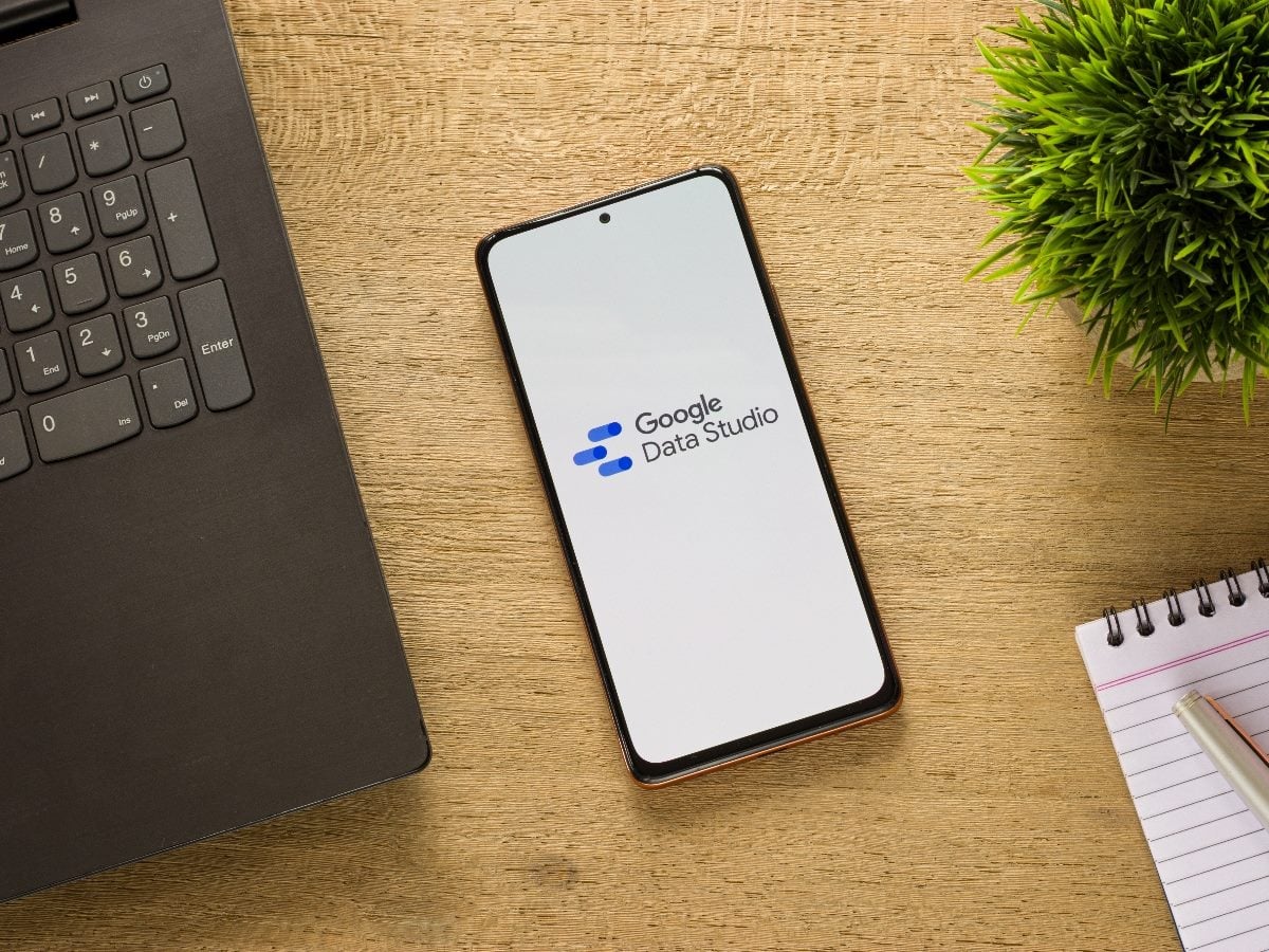 Smartphone displaying the Google Data Studio logo on a desk beside a laptop, notepad and plant, representing modern analytics and data visualization tools.