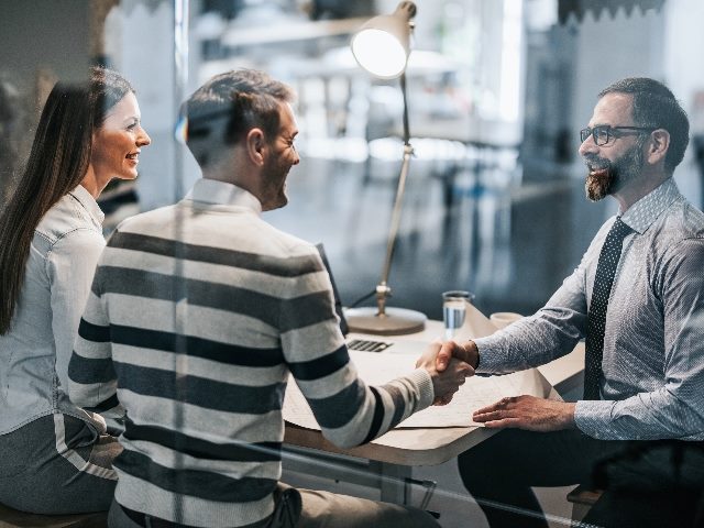 A man and woman sitting at a table across another man. The men are shaking hands. All have smiles.