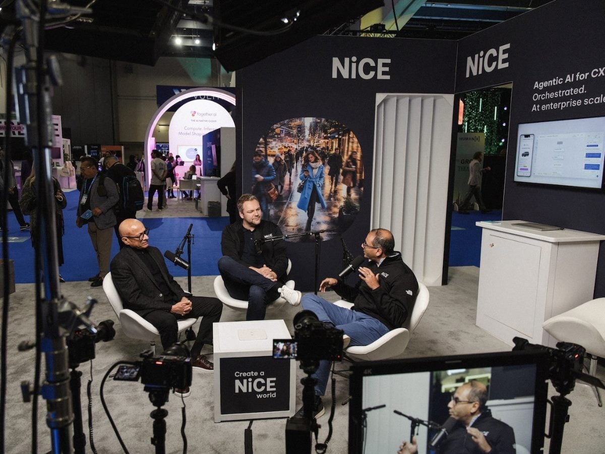 Three individuals seated in white chairs conduct a recorded interview at the NiCE booth on a conference expo floor, with microphones, cameras, and studio lighting in frame. NiCE branding and the tagline "Agentic AI for CX. Orchestrated. At enterprise scale." are visible on the booth backdrop.