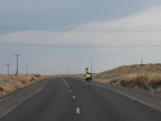 Two bikers ride forward in an otherwise empty road.