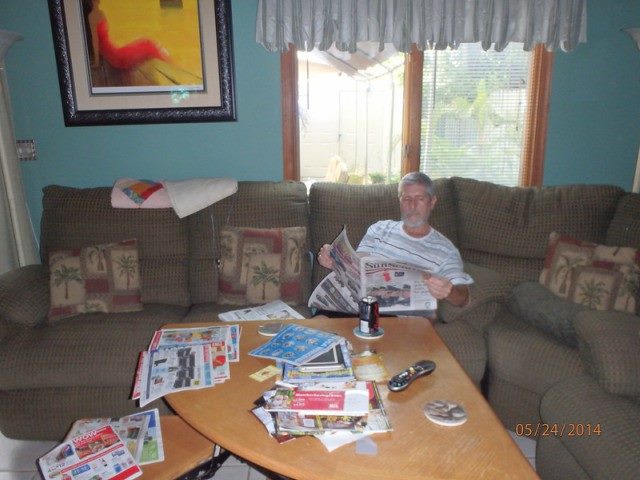 A man sitting on a couch reading a newspaper with other newspaper sections strewn across a coffee table.