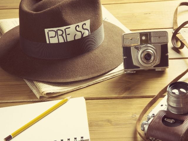 hat with the label "press" on it, a notebook with a pencil on it and two cameras spread across a table.