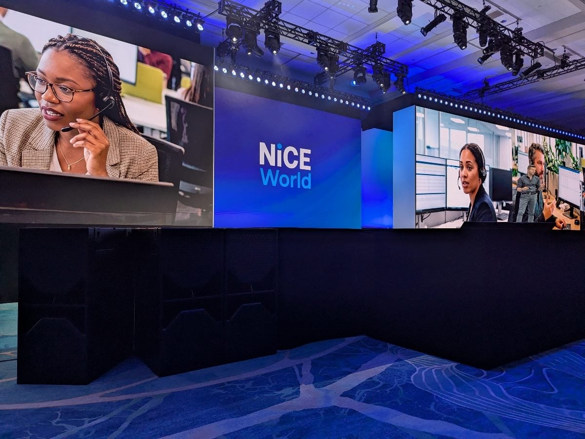 The NiCE World 2026 main stage at the Walt Disney Dolphin Resort in Orlando, featuring large LED screens displaying contact center agents at work, flanking the illuminated NiCE World logo. Philipp Heltewig, Chief AI Officer at NiCE Cognigy, is visible on the right screen delivering his keynote address.