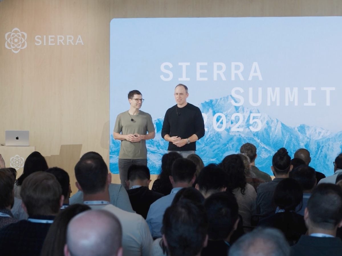 Two presenters stand on stage addressing an audience at Sierra Summit 2025, with a large screen behind them displaying “Sierra Summit 2025” over a mountain backdrop and Sierra branding visible on the stage wall.