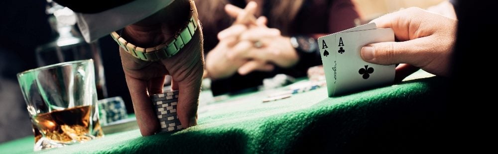 People sit at a green covered table playing poker with one person lifting two cards that are Aces, a drink in a rocks glass is visible on the left and a person's hand holding poker chips is next to it in piece about the skills required for using AI in marketing effectively.