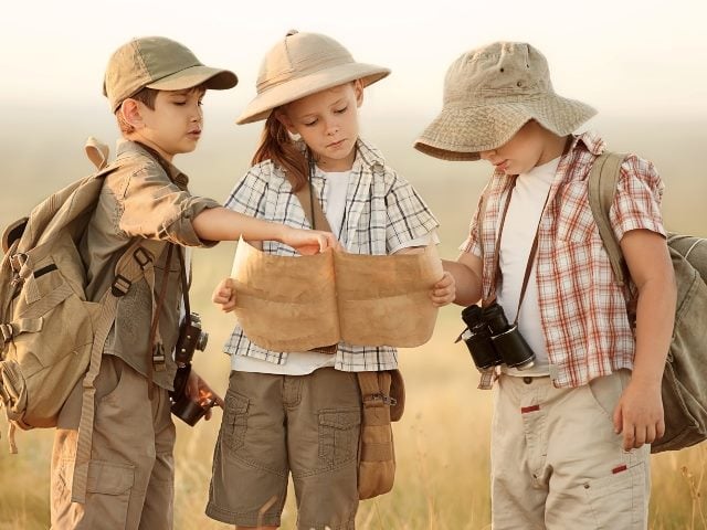 A group of three kids dressed in safari outfits look at a map at sunset representing undertaking the journey of customer journey mapping. 