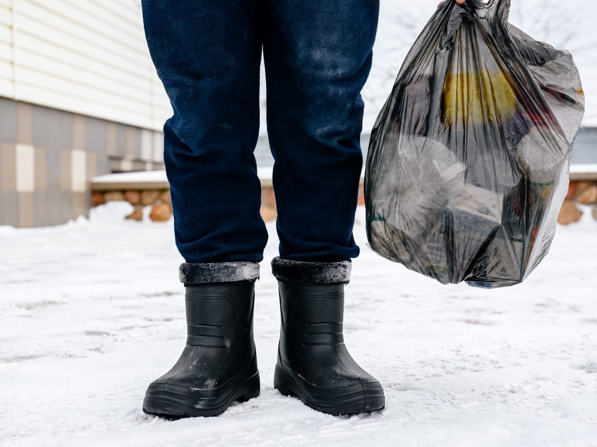 Person standing outdoors in snowy conditions, wearing dark winter boots and pants, holding a black plastic trash bag filled with household waste.