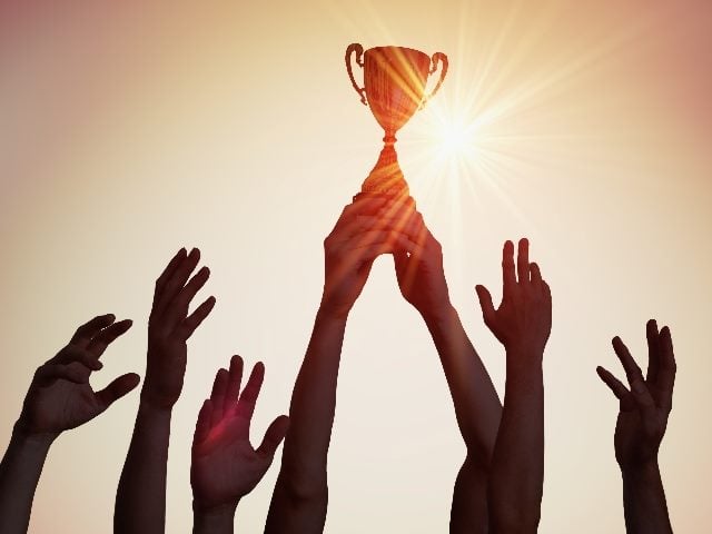 A winning teams hands hold their trophy up in  the air with the sun in the background indicating the importance of customer success and retention. 