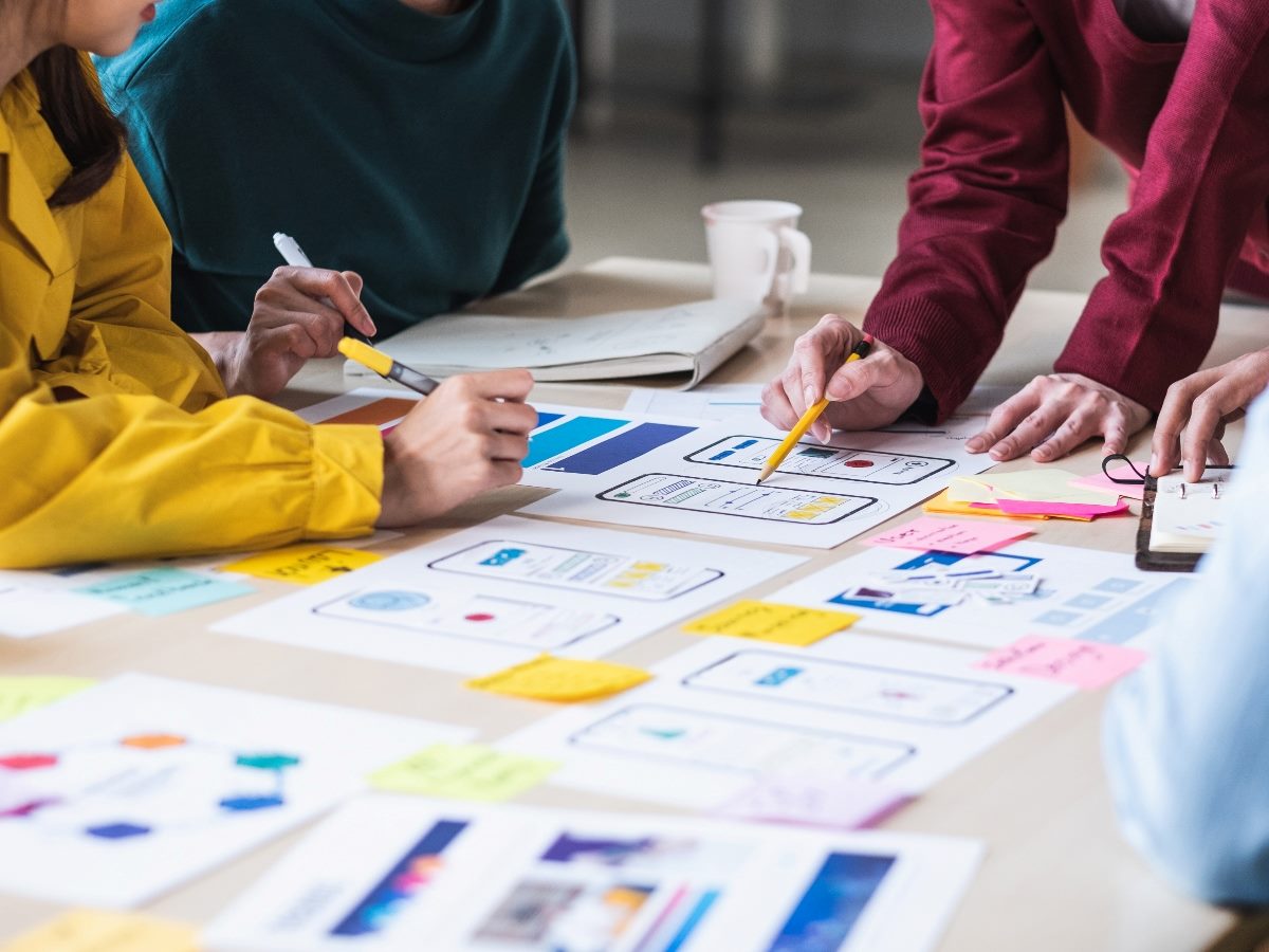 Team of designers collaborating on user experience layouts, reviewing wireframes and colorful sticky notes spread across a table during a design planning session.