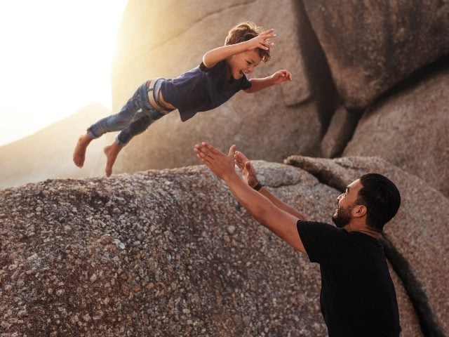 A young boy jumps from a large rock on a beach into the arms of an adult male standing waiting to catch him with golden sunshine in the background, suggesting the importance of trust in CX and EX.