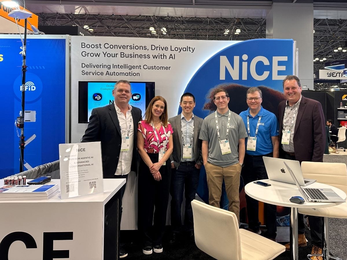 NiCE team members stand at the NiCE booth at NRF 2026, posing in front of a large blue NiCE backdrop promoting AI-powered customer service automation, with demo screens, signage and a small meeting table visible on the show floor.