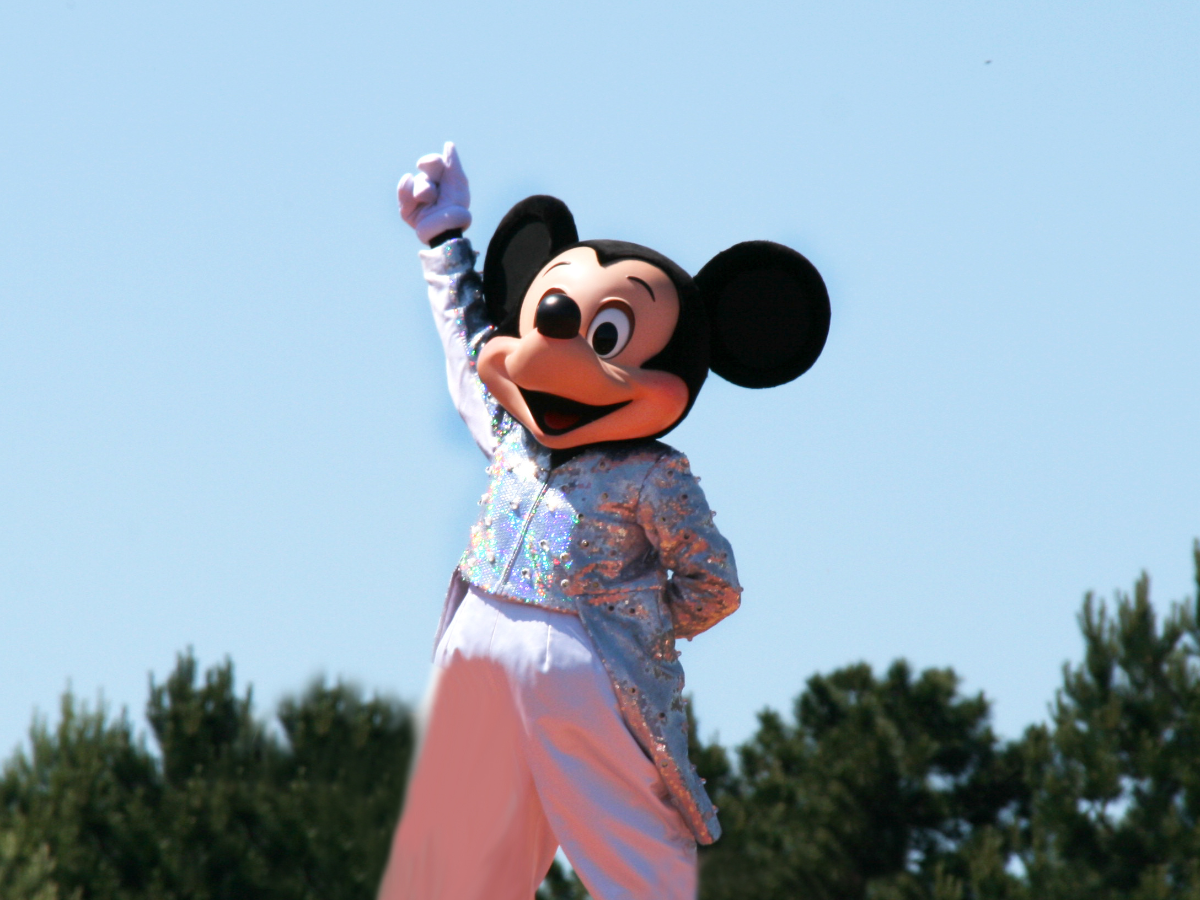 Mickey Mouse character in a sparkling silver jacket and white pants, raising one arm in a celebratory pose outdoors under a clear blue sky.