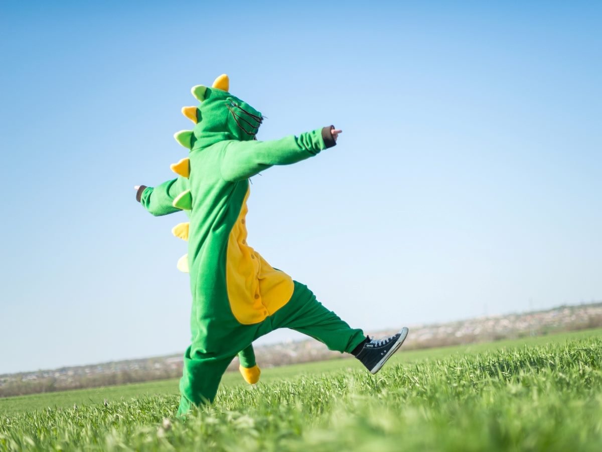 Person in a green dinosaur costume walking through a grassy field with arms outstretched.