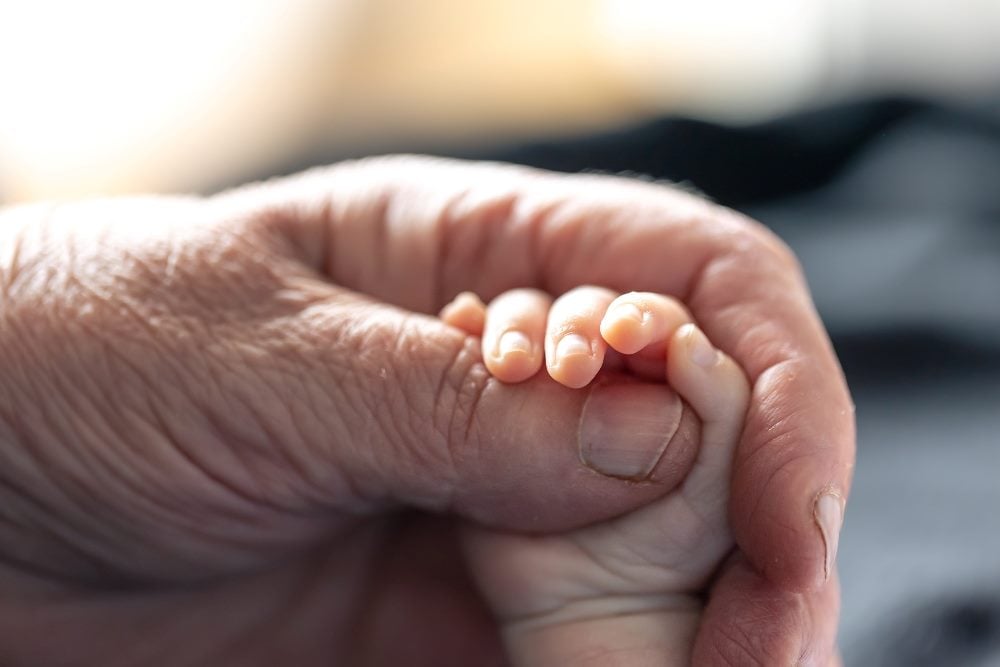 The hand of a senior person delicately holds the new hand of a baby, indicating the need for the human touch in AI customer experience.
