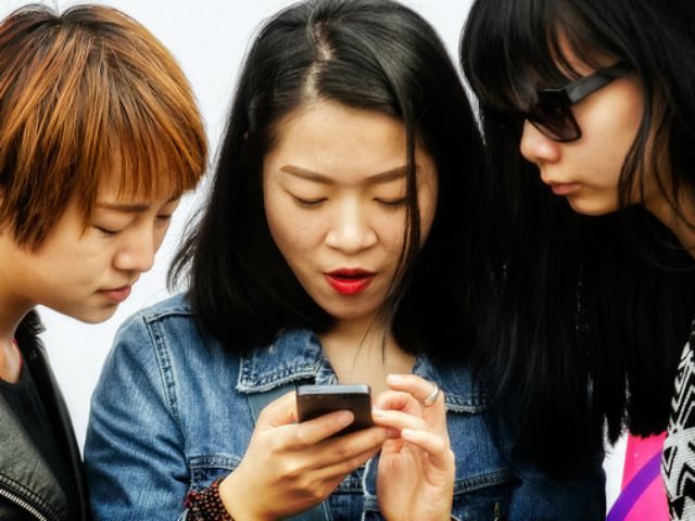 two girls look over shoulder of third girl on her phone