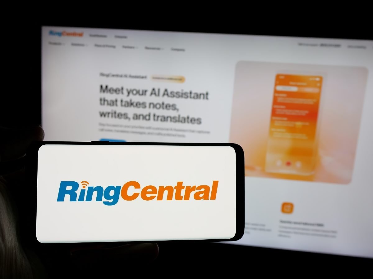 A smartphone displays the RingCentral logo in front of a blurred computer screen showing RingCentral’s AI assistant webpage.