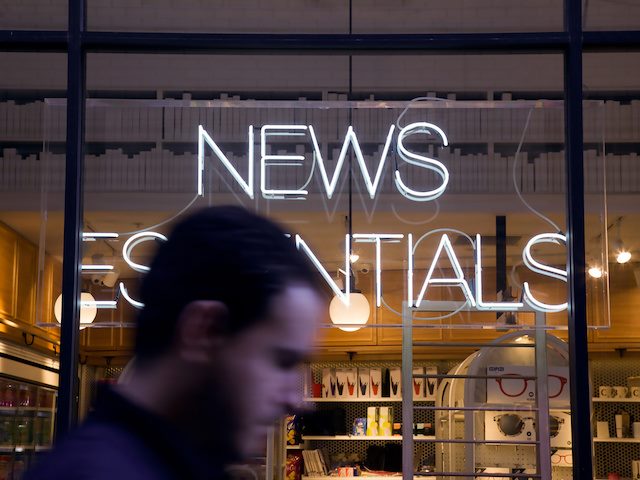 man walking in front of a "news essentials" store with neon sign