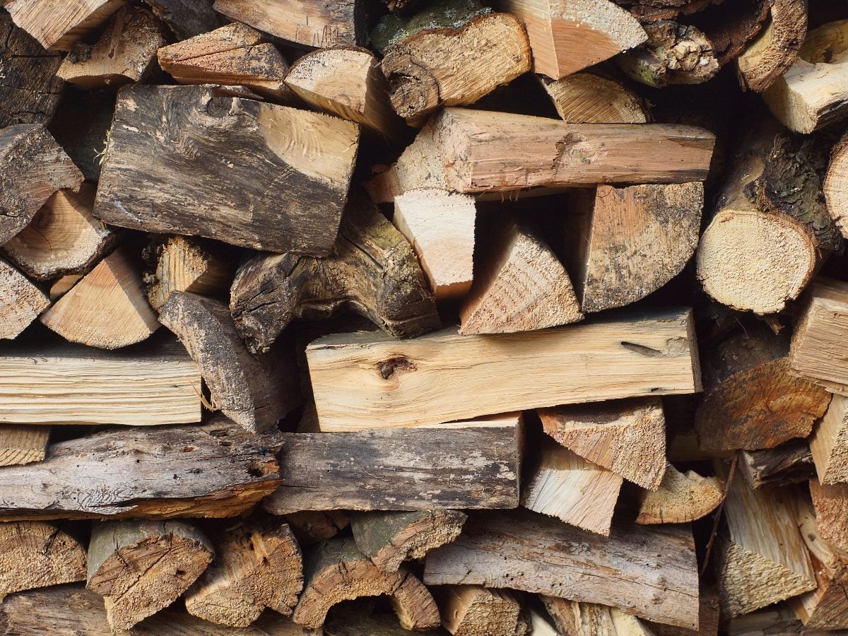 Close-up image of a stacked pile of split firewood logs in various sizes and shapes, showing rough bark textures, exposed wood grain and natural brown tones.