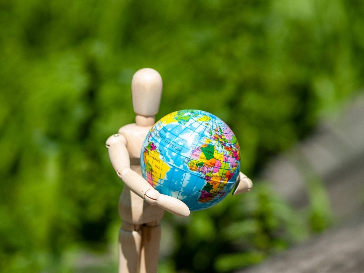 Wooden artist mannequin holding a colorful globe outdoors against a blurred green background, symbolizing humanity, global systems or human-centered technology.