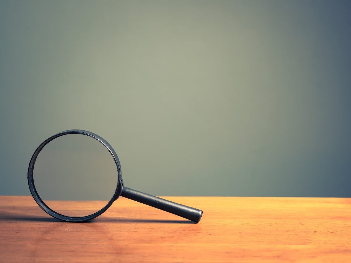 A black magnifying glass resting on a wooden surface against a soft green background, symbolizing close inspection, visibility and deeper analysis.