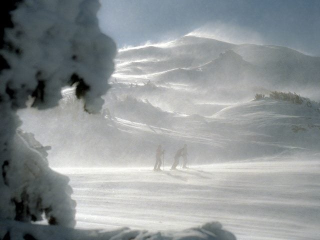 Skiers in the distance walking in a windy snowy scene in the mountains.